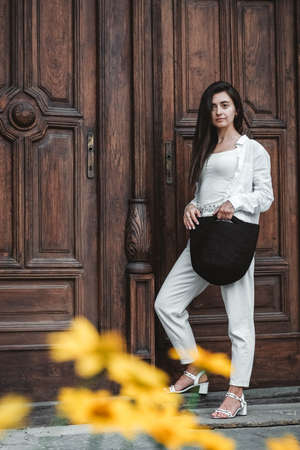 Beautiful young brunette woman wearing in white clothes, holding knitted bag on the background of old wooden doors. Copy, empty space for text.の写真素材