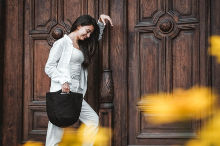 Beautiful young brunette woman wearing in white clothes, holding knitted bag on the background of old wooden doors. Copy, empty space for text.の写真素材
