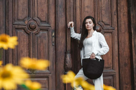 Beautiful young brunette woman wearing in white clothes, holding knitted bag on the background of old wooden doors. Copy, empty space for text.の写真素材