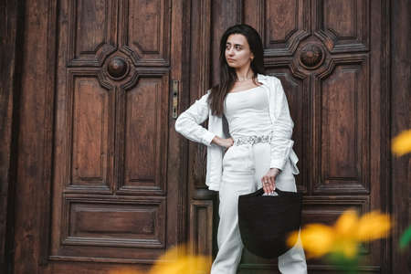 Beautiful young brunette woman wearing in white clothes, holding knitted bag on the background of old wooden doors. Copy, empty space for text.の写真素材
