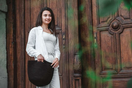 Beautiful young brunette woman wearing in white clothes, holding knitted bag on the background of old wooden doors. Copy, empty space for text.の写真素材