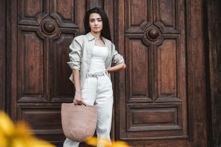 Beautiful young brunette woman wearing in white clothes, holding knitted bag on the background of old wooden doors. Copy, empty space for text.の写真素材