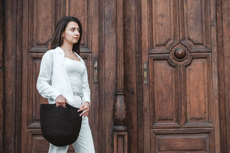 Beautiful young brunette woman wearing in white clothes, holding knitted bag on the background of old wooden doors. Copy, empty space for text.の写真素材