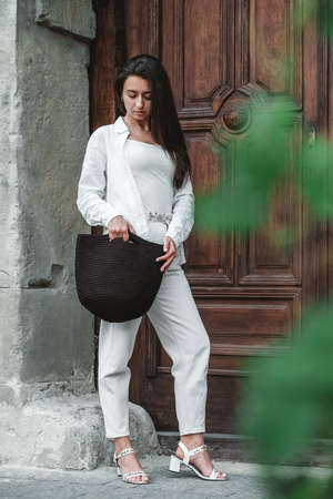 Beautiful young brunette woman wearing in white clothes, holding knitted bag on the background of old wooden doors. Copy, empty space for text.の写真素材