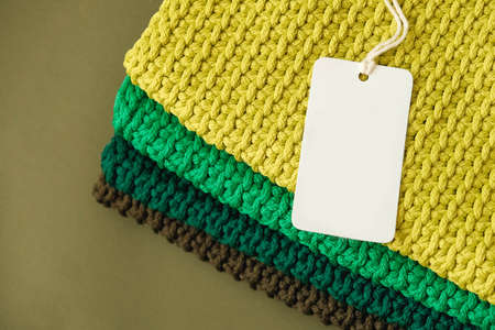 Stack of knitted material from threads of yellow, green, brown colors with blank Price Tag on a blue background. Top view. Copy, empty space for text.の写真素材
