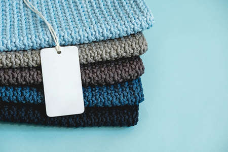 Stack of knitted material from threads of dark blue, light blue, gray colors with blank Price Tag on a blue background. Copy, empty space for text.の写真素材