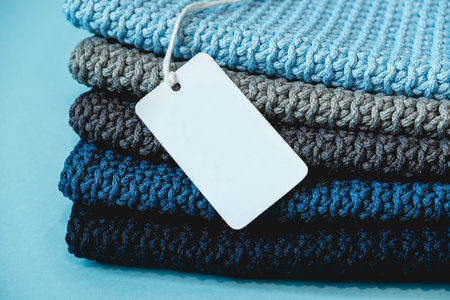 Stack of knitted material from threads of dark blue, light blue, gray colors with blank Price Tag on a blue background. Copy, empty space for text.の写真素材