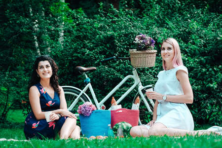 Two young beautiful women in trendy summer sundress sitting on a plaid in the park on the background of a bicycle. Positive models sitting on plaid on grass, drink juice and eat fruit. Carefree women making picnic outside. Copy, empty space for text.の写真素材