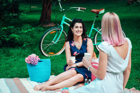 Two young beautiful women in trendy summer sundress sitting on a plaid in the park on the background of a bicycle. Positive models sitting on plaid on grass, drink juice and eat fruit. Carefree women making picnic outside. Copy, empty space for text.の写真素材