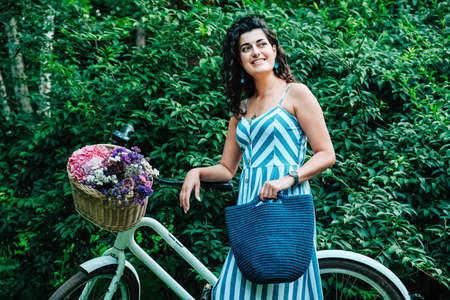 Young beautiful woman dressed in a in a striped dress is standing next to a bicycle with a basket of flowers in the park. Copy, empty space for text.の写真素材