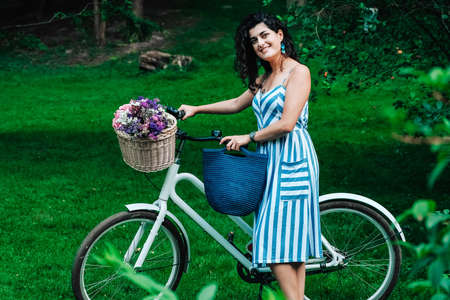 Young beautiful woman dressed in a in a striped dress is standing next to a bicycle with a basket of flowers in the park. Copy, empty space for text.の写真素材