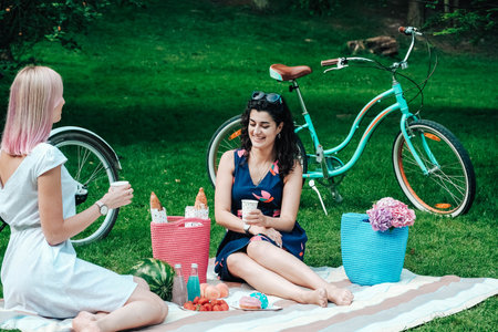 Two young beautiful women in trendy summer sundress sitting on a plaid in the park on the background of a bicycle. Positive models sitting on plaid on grass, drink juice and eat fruit. Carefree women making picnic outside. Copy, empty space for text.の写真素材