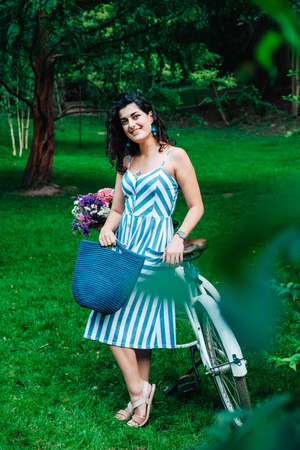 Young beautiful woman dressed in a in a striped dress is standing next to a bicycle with a basket of flowers in the park. Copy, empty space for text.の写真素材
