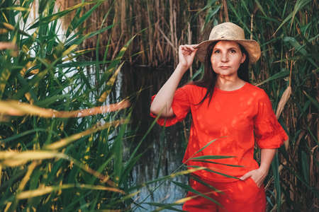 Portrait of a beautiful woman dressed in red clothes standing against the backdrop of reeds and lake. Copy, empty space for text.の写真素材