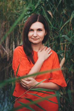 Portrait of a beautiful woman dressed in red clothes standing against the backdrop of reeds and lake. Copy, empty space for text.の写真素材