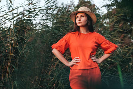 Portrait of a beautiful woman in a straw hat dressed in red clothes on a background of reeds and lake. Copy, empty space for text.の写真素材