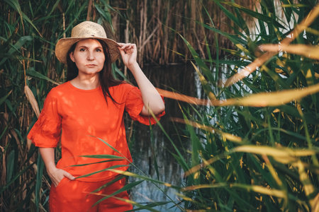 Portrait of a beautiful woman in a straw hat dressed in red clothes on a background of reeds and lake. Copy, empty space for text.の写真素材