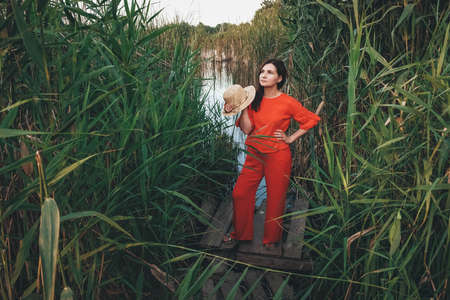 Portrait of a beautiful woman in a straw hat dressed in red clothes on a background of reeds and lake. Copy, empty space for text.の写真素材