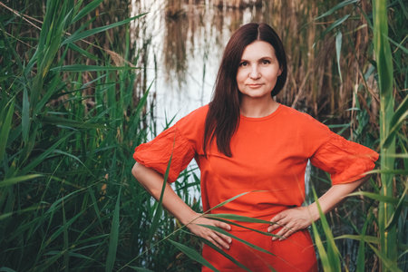 Portrait of a beautiful woman in a straw hat dressed in red clothes on a background of reeds and lake. Copy, empty space for text.の写真素材