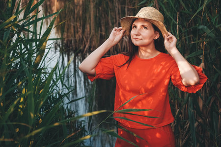 Beautiful woman in a straw hat dressed in red clothes on a background of reeds and lake. Copy, empty space for text.の写真素材