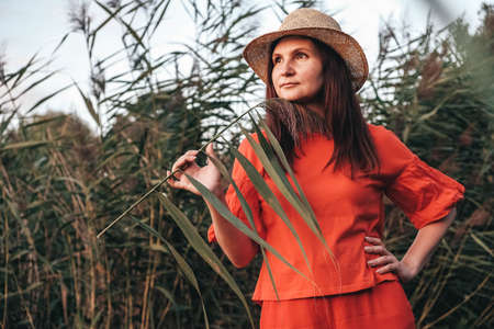 Beautiful woman in a straw hat dressed in red clothes on a background of reeds and lake. Copy, empty space for text.の写真素材