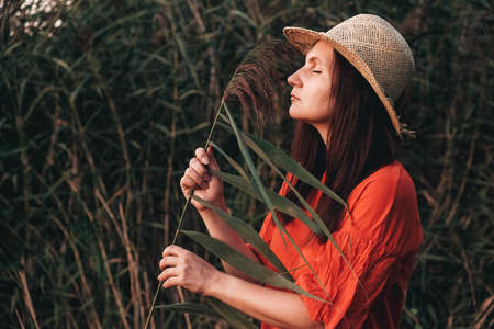 Beautiful woman in a straw hat dressed in red clothes on a background of reeds and lake. Copy, empty space for text.の写真素材
