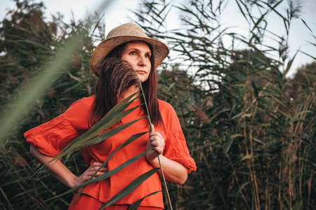 Beautiful woman in a straw hat dressed in red clothes on a background of reeds and lake. Copy, empty space for text.の写真素材