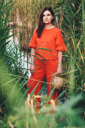 Beautiful woman in a straw hat dressed in red clothes on a background of reeds and lake. Copy, empty space for text.の写真素材