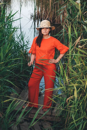 Beautiful woman in a straw hat dressed in red clothes on a background of reeds and lake. Copy, empty space for text.の写真素材