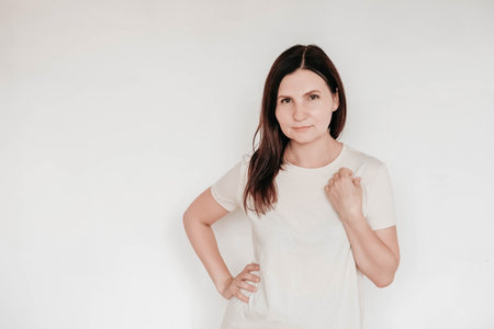 Beautiful brunette woman keeps hands on cheek, dressed in casual white t shirt, has minimal makeup, poses indoor on a white background. Copy, empty space for text.の写真素材