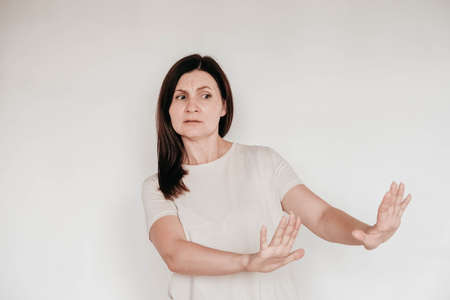 Beautiful brunette woman keeps hands on cheek, dressed in casual white t shirt, has minimal makeup, poses indoor on a white background. Copy, empty space for text.の写真素材
