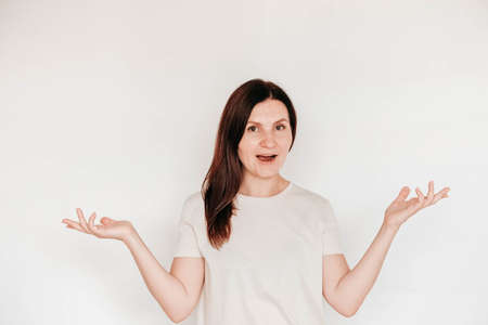 Beautiful brunette woman keeps hands on cheek, dressed in casual white t shirt, has minimal makeup, poses indoor on a white background. Copy, empty space for text.の写真素材