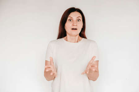 Beautiful brunette woman keeps hands on cheek, dressed in casual white t shirt, has minimal makeup, poses indoor on a white background. Copy, empty space for text.の写真素材
