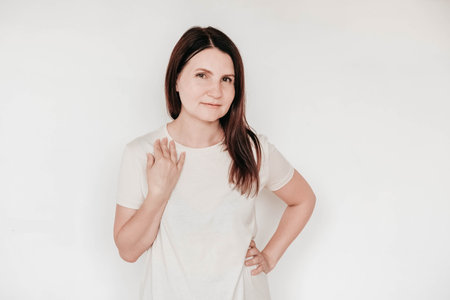 Beautiful brunette woman keeps hands on cheek, dressed in casual white t shirt, has minimal makeup, poses indoor on a white background. Copy, empty space for text.の写真素材