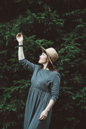 Beautiful red-haired young woman in a straw hat and dress on a background of green forest and trees. Place for text or advertising.の写真素材