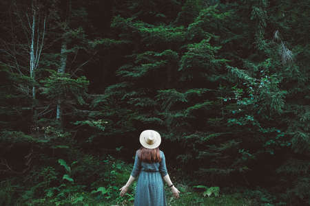Beautiful red-haired young woman in a straw hat and dress on a background of green forest and trees. Place for text or advertising.の写真素材