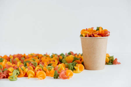 Multicolored pasta in a disposable kraft paper cup scattered on a white background. Copy, empty space for text.の写真素材