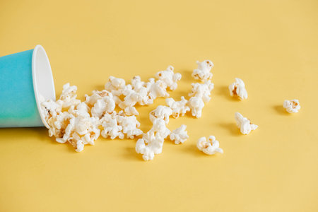 Popcorn in a blue paper cup scattered on a yellow background. Minimalist concept. Copy, empty space for text.の写真素材