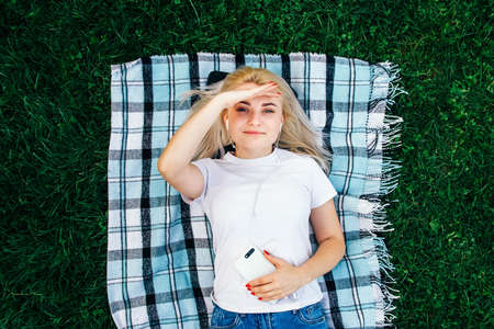 Beautiful blonde woman in headphones and with a smartphone in his hands look at the device sitting on grass outdoors in park. Place for text or advertising.の写真素材