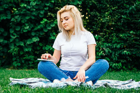 Beautiful blonde woman in headphones and with a smartphone in his hands look at the device sitting on grass outdoors in park. Place for text or advertising.の写真素材