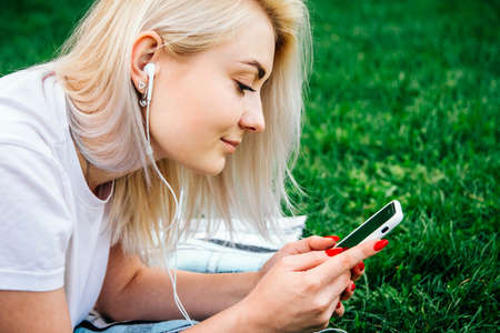 Beautiful blonde woman in headphones and with a smartphone in his hands look at the device sitting on grass outdoors in park. Place for text or advertising.の写真素材