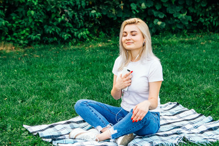 Beautiful blonde woman in headphones and with a smartphone in his hands look at the device sitting on grass outdoors in park. Place for text or advertising.の写真素材