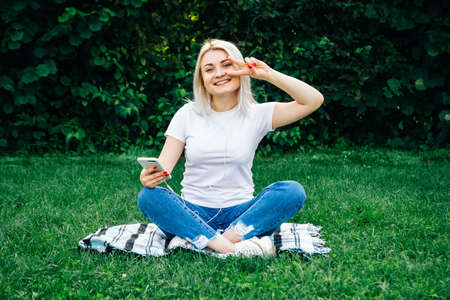 Beautiful blonde woman in headphones and with a smartphone in his hands look at the device sitting on grass outdoors in park. Place for text or advertising.の写真素材
