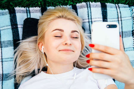 Beautiful blonde woman in headphones and with a smartphone in his hands look at the device sitting on grass outdoors in park. Place for text or advertising.の写真素材