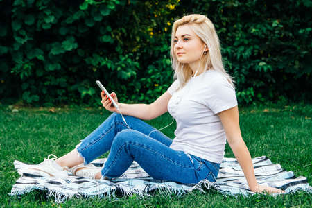 Beautiful blonde woman in headphones and with a smartphone in his hands look at the device sitting on grass outdoors in park. Place for text or advertising.の写真素材
