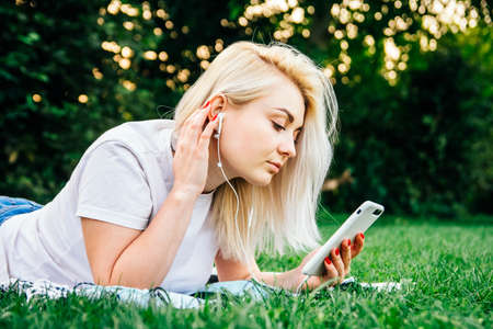 Beautiful blonde woman in headphones and with a smartphone in his hands look at the device sitting on grass outdoors in park. Place for text or advertising.の写真素材