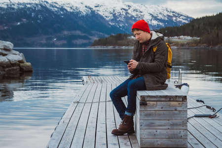 Traveler man with a yellow backpack wearing sits near the wooden red colored house. Travel lifestyle conceptの写真素材