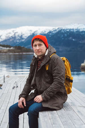 Traveler man with a yellow backpack wearing sits near the wooden red colored house. Travel lifestyle conceptの写真素材
