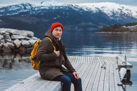 Traveler man with a yellow backpack wearing sits near the wooden red colored house. Travel lifestyle conceptの写真素材