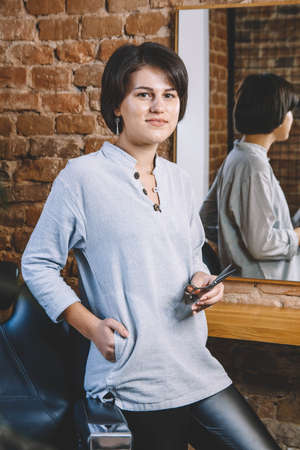 Young beautiful hairdresser woman with scissors in hand against a brick wall background. Barbershop advertising concept. Place for text or advertising.の写真素材